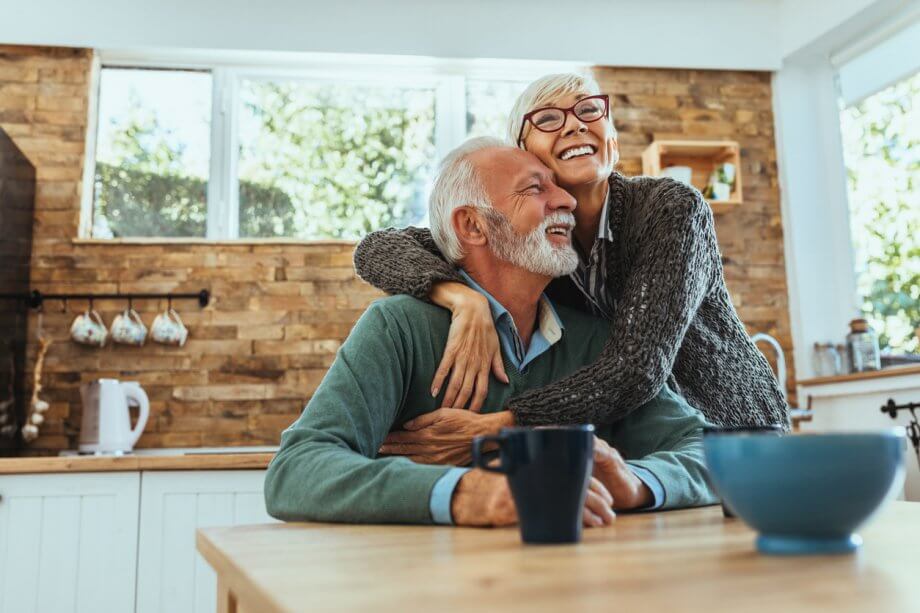 Older couple holding each other and smiling while sitting in a kitchen.