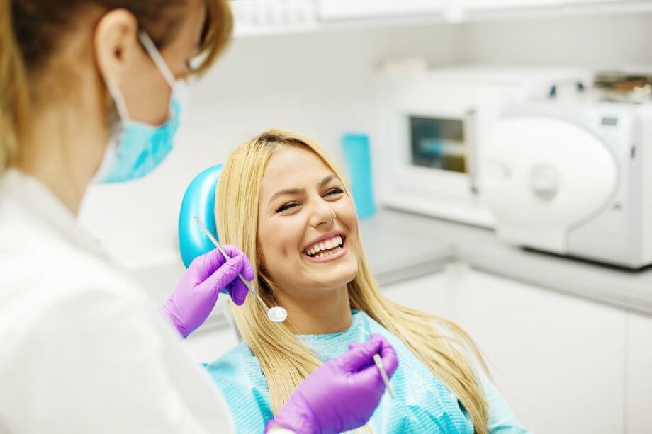 Blonde woman sitting in dentist chair smiling and laughing while a female dental hygienist sits next to her.