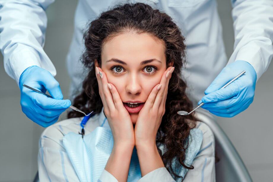 Woman with long curly brown hair scared of the dentist as the dentist stands behind her holding tools.