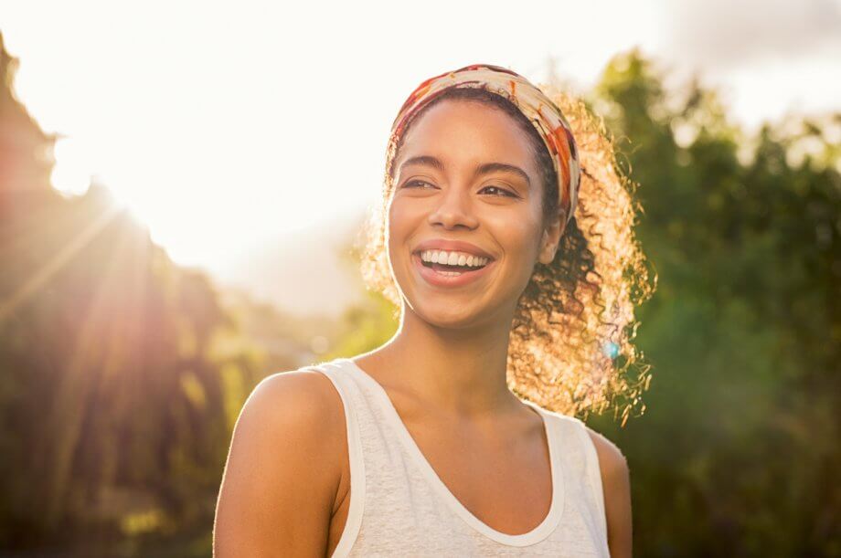 Woman smiling wearing a tank top in nature with the sun setting behind her.