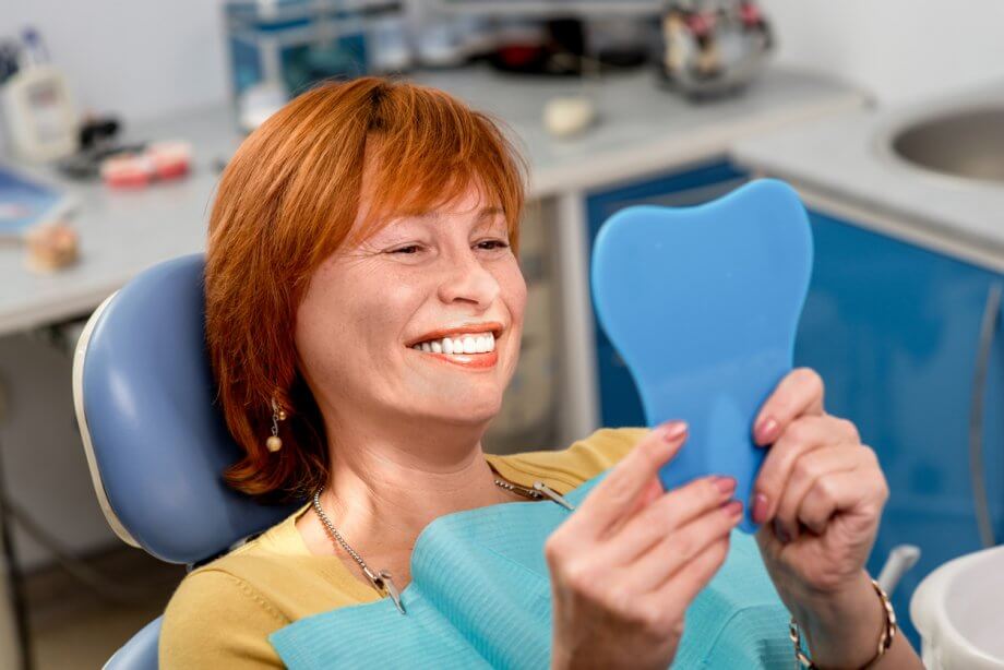 Photograph of a woman with short, straight red hair smiling while looking in a handheld mirror while at the dentist.